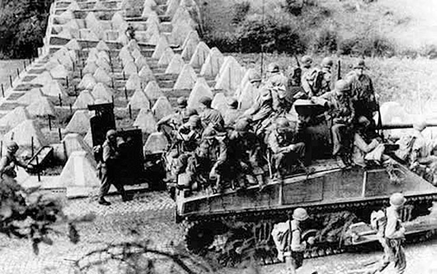An M4 Sherman tank carries Third U.S. Army troops through the dragon&rsquo;s teeth antitank barriers of the Siegfried Line.