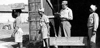 4. Sergeant Harold “Bud” Banker checks a British Bren light machinegun out of the Detachment 101 supply warehouse in Nazira, India. The Indian porter in the front carries a case of .303 caliber bullets on his head.