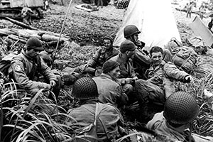 Men of the First Regiment command group huddle on Beach 9-Blue on D-Day 15 August 1943. The white triangular shape behind the men is a beach marking panel erected to guide the subsequent waves of troops to the beach.